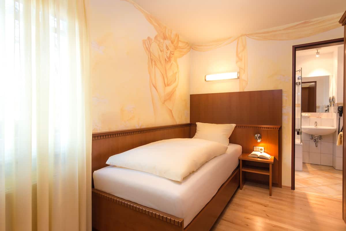 A hotel room with a bed, bedside table, lamp, and a bathroom visible through an open door.