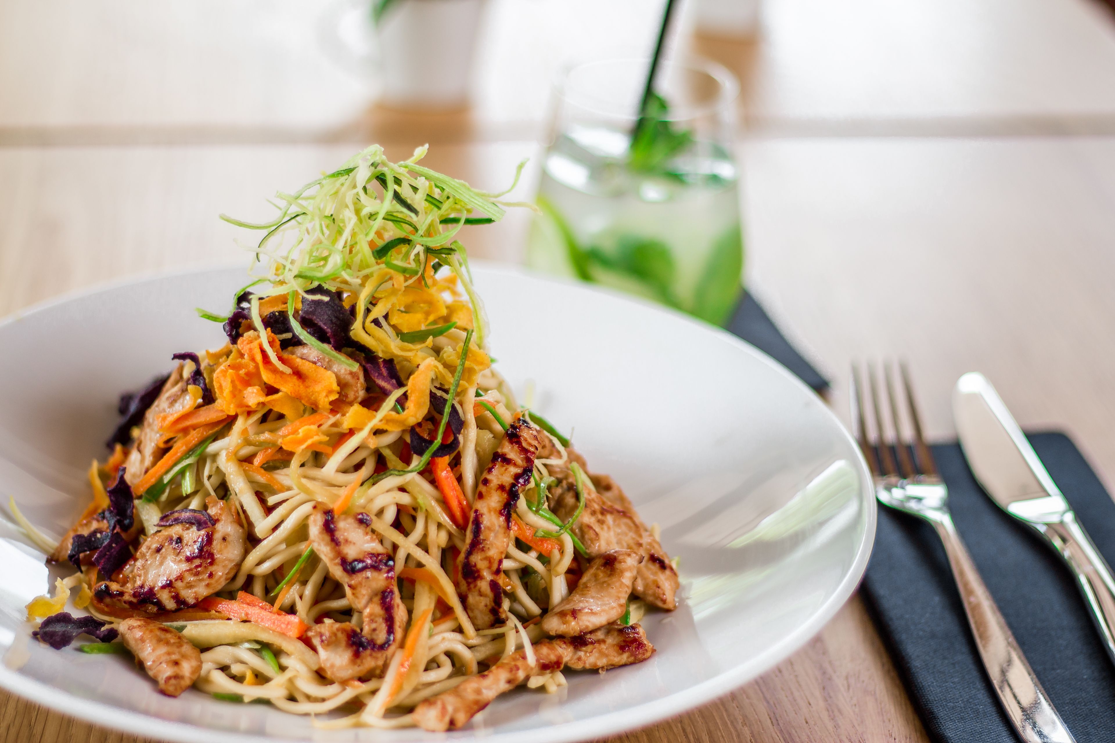 A plate of stir-fried noodles with vegetables and chicken, accompanied by a glass of iced tea.