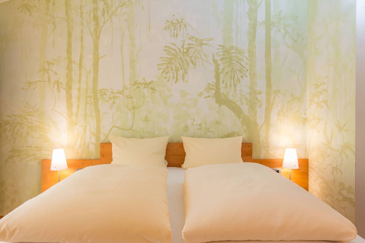 A double bed with two pillows and two bedside lamps, set against a wall with a tropical wallpaper.