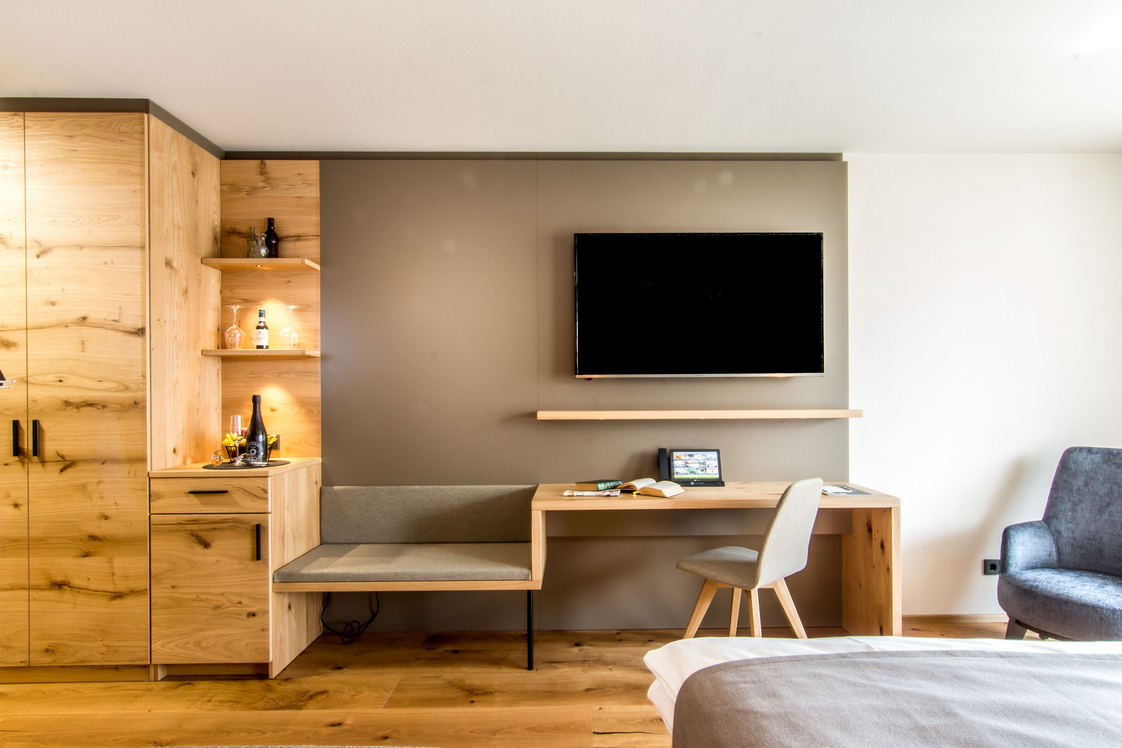 A modern bedroom with a wooden desk, a flat-screen TV, a grey armchair, and a bed.