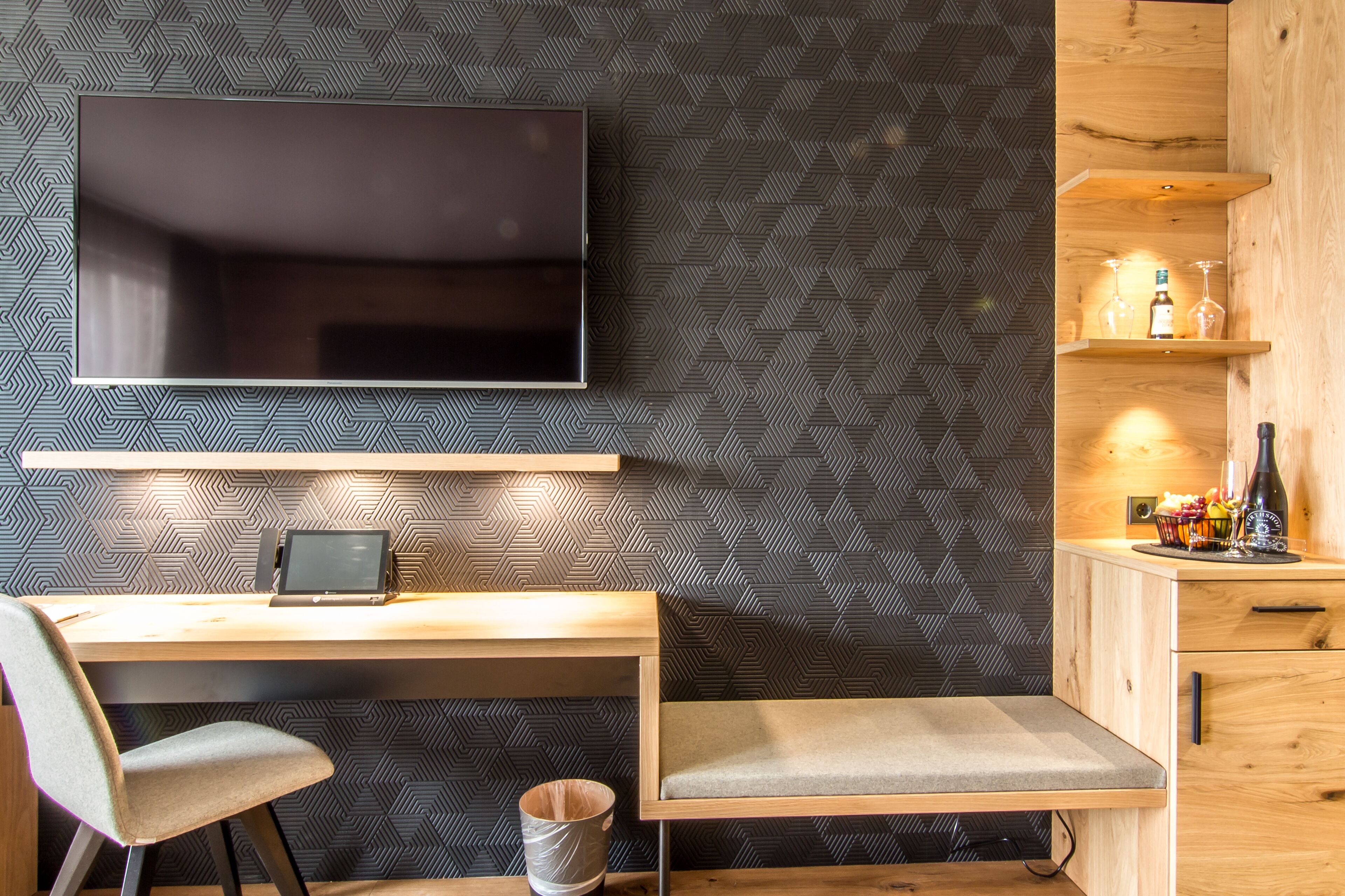 Una habitación moderna con una televisión de pantalla plana montada en la pared, un escritorio de madera con una silla, un cubo de basura pequeño y una estantería con botellas y vasos.