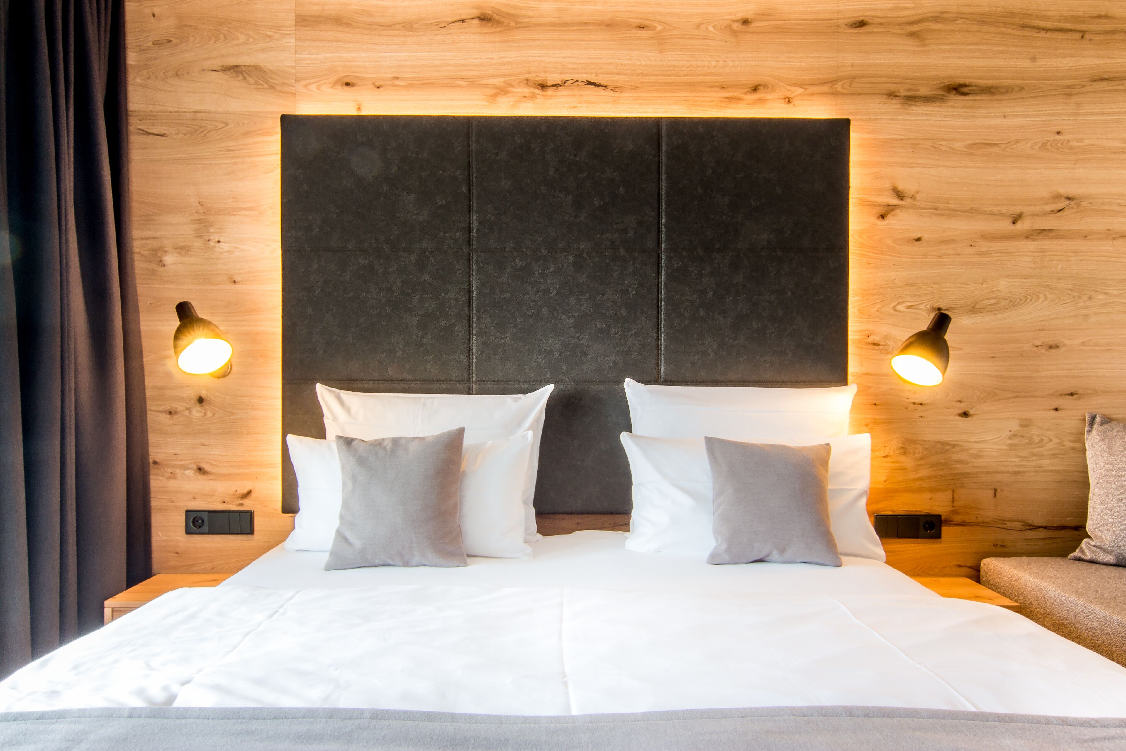 A neatly made bed with white linens and two pillows, a wooden headboard, and two wall-mounted lamps.