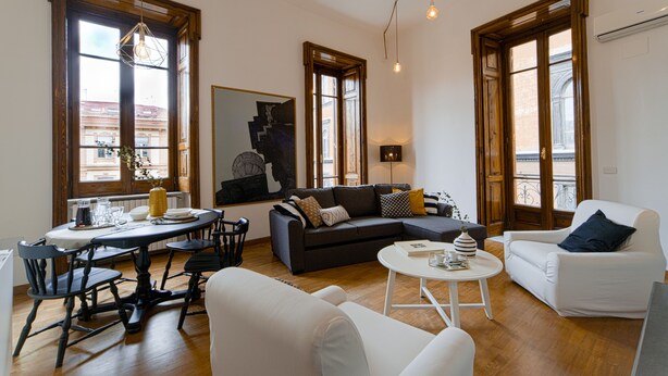 A spacious living room with a dark-colored sofa, a white armchair, and a dining area with a table and chairs. - Casa Frangiò - Naples Historical Centre (Naples)