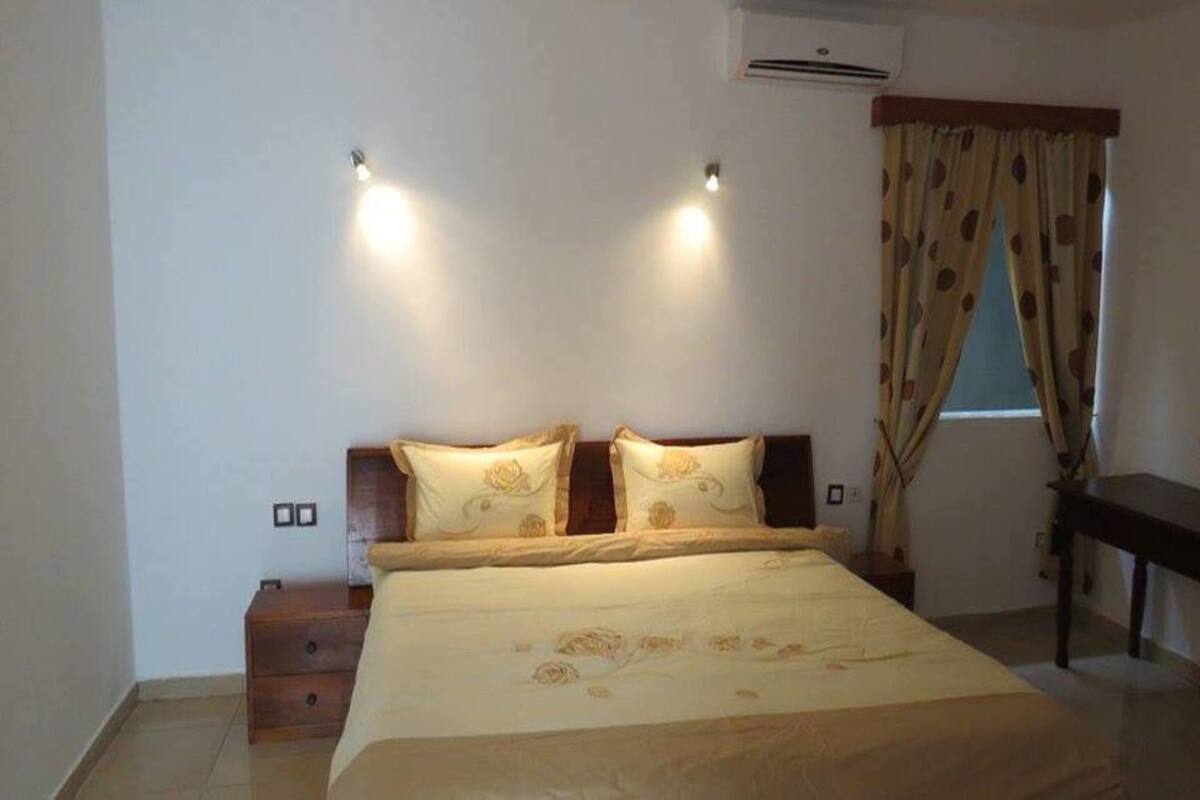 A bedroom with a bed, two bedside tables, a nightstand, a window with curtains, and an air conditioning unit.