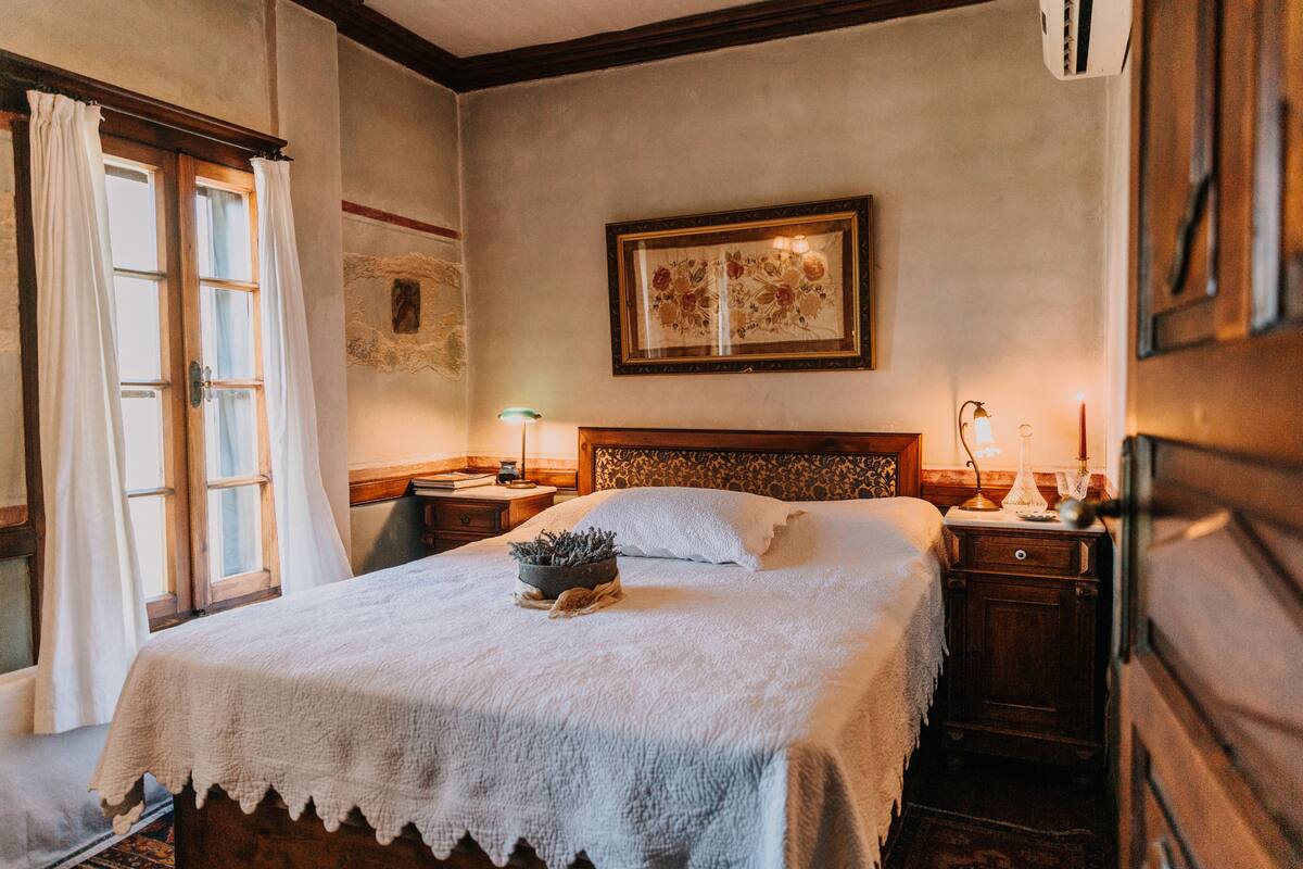 A bedroom with a large bed, wooden headboard, bedside tables, a nightstand, a framed floral artwork, and a window with curtains.