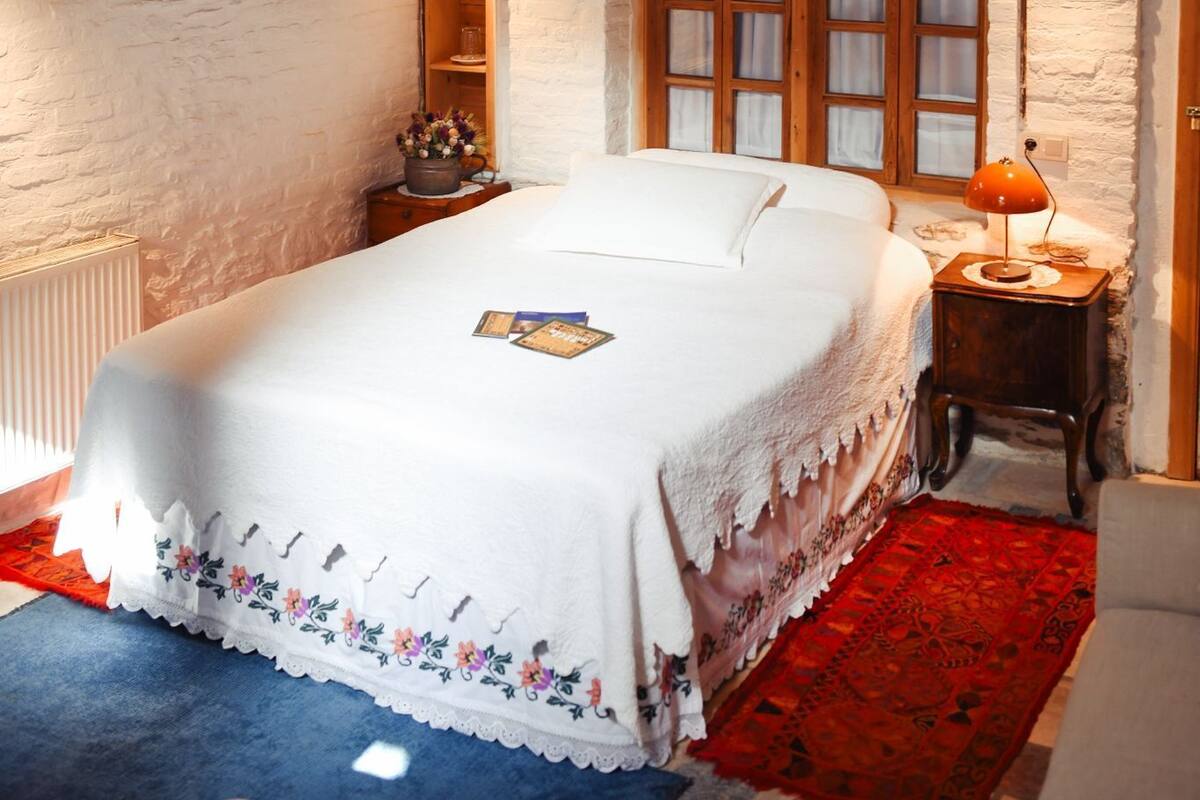 A cozy bedroom with a wooden ceiling, a large window with curtains, a bedside table with a lamp, and a red rug.