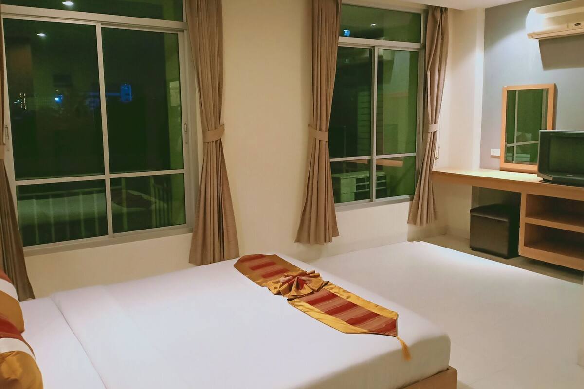 A hotel room with a bed, a desk with a television, and large windows with curtains.