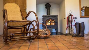 Una habitación con una silla de madera, una silla tejida, una chimenea con leña y un perchero con botas.