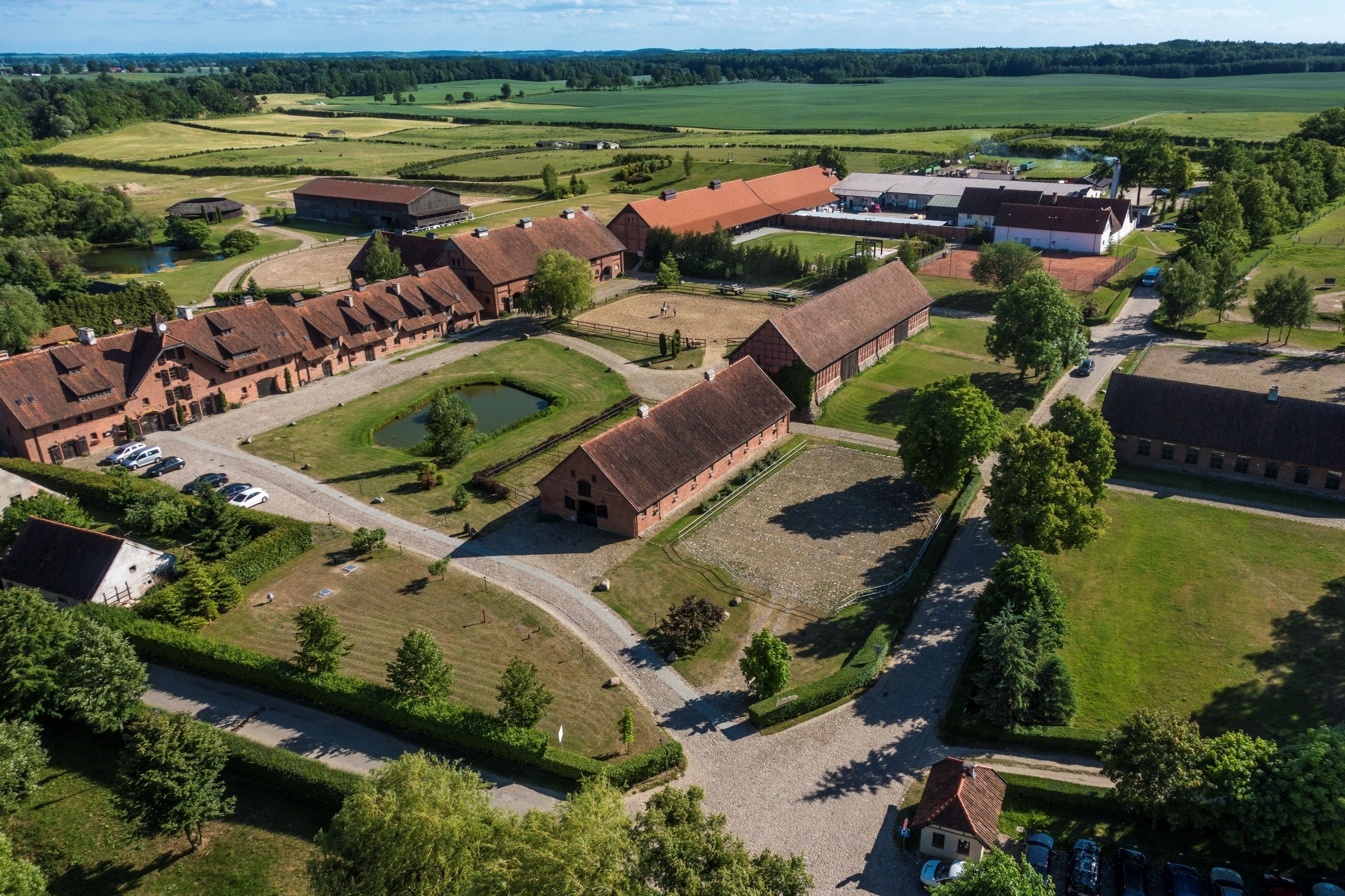 An aerial view of a rural estate with multiple buildings, including barns and a large house, surrounded by green fields and a small pond.