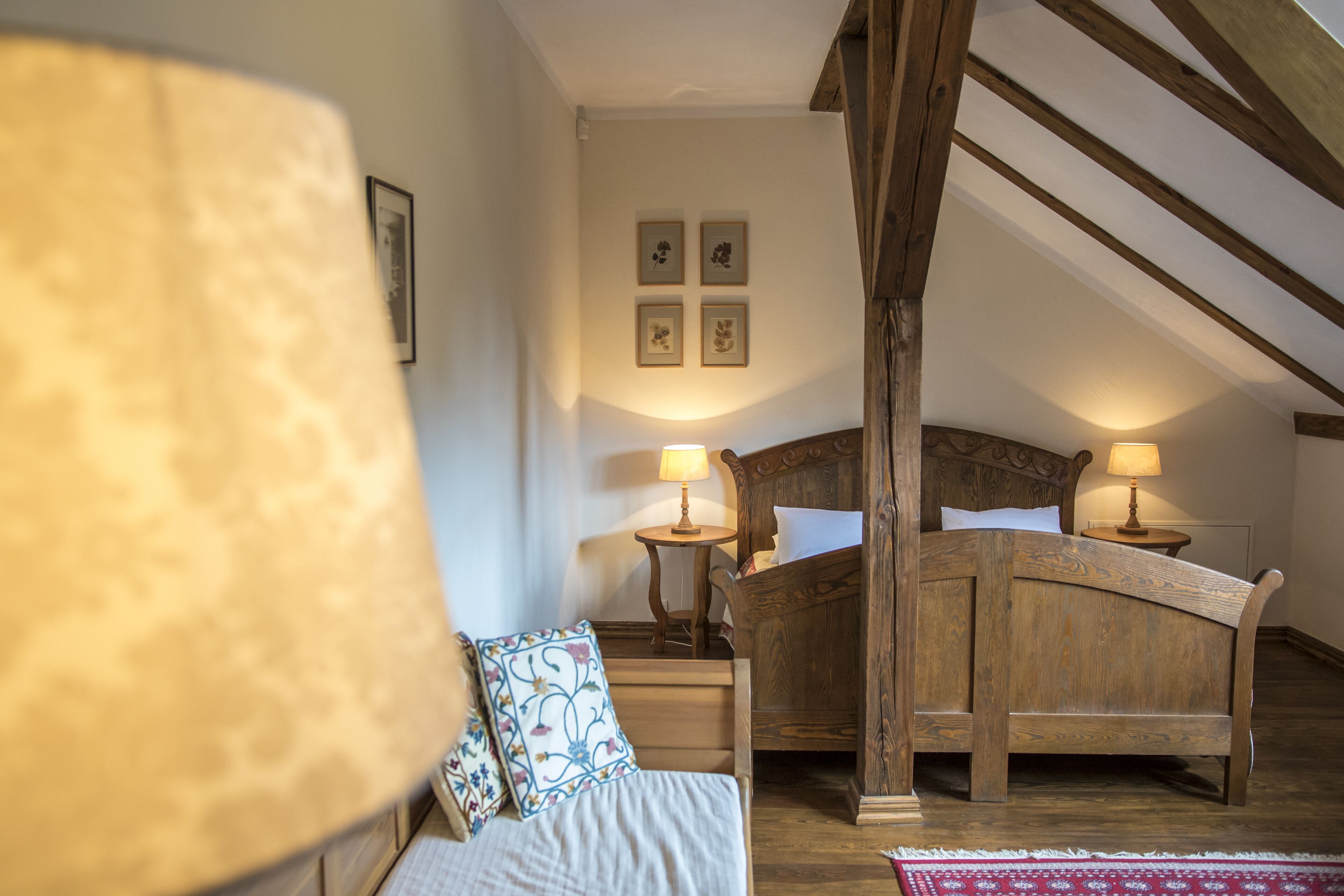 A bedroom with a wooden bed, two bedside tables with lamps, a red patterned rug, and four framed pictures on the wall.