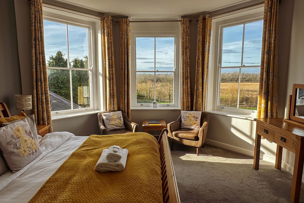 Ein Schlafzimmer mit einem großen Bett, zwei Sesseln, einem kleinen Tisch und einem Schreibtisch. Drei Fenster bieten Ausblick ins Freie.