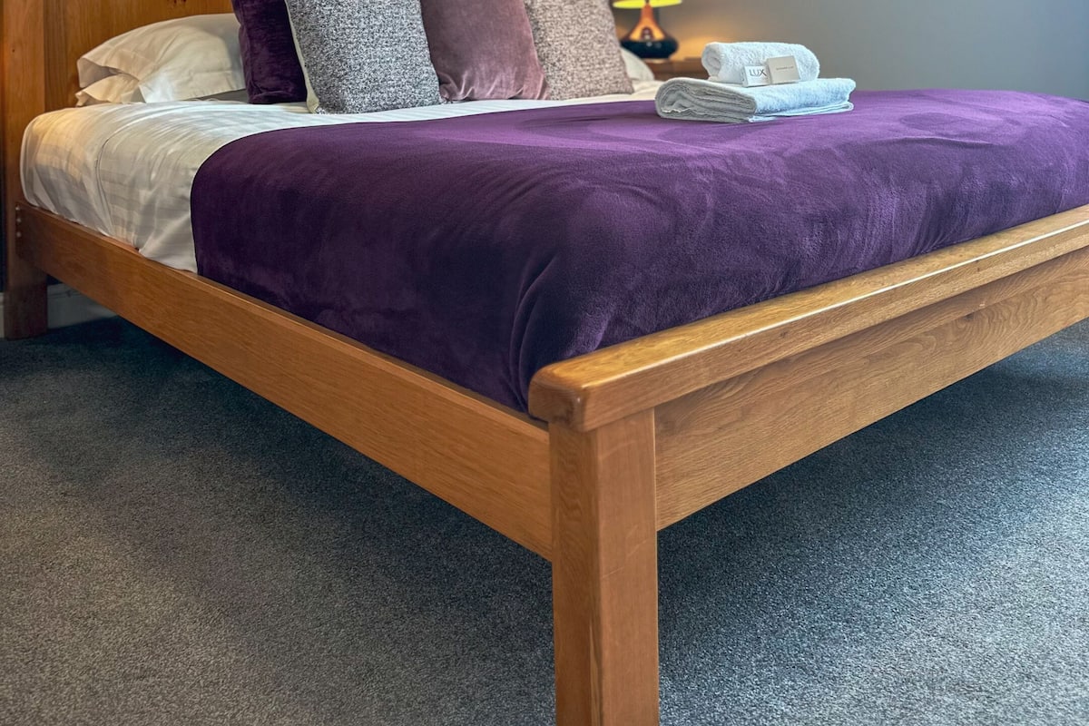 Ein ordentlich bezogenes Bett mit einer violetten Decke, einem hölzernen Kopfteil und einem Kissen mit floralem Muster.