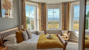 Ein Schlafzimmer mit einem großen Bett, zwei Nachttischen, einem Fenster mit Vorhängen und Blick ins Freie.
