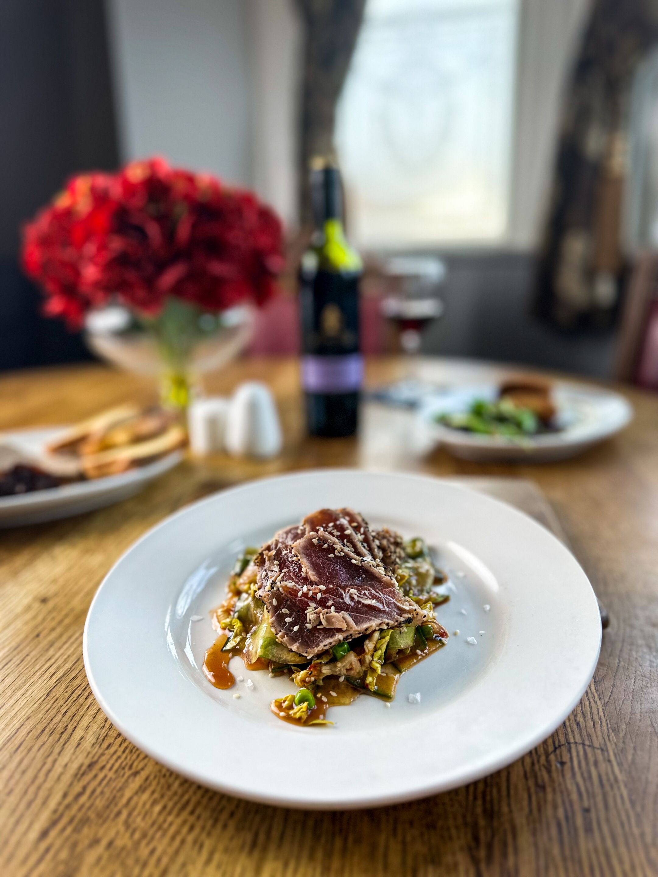 Una mesa de comedor con un plato de comida, una botella de vino y un jarrón de flores.