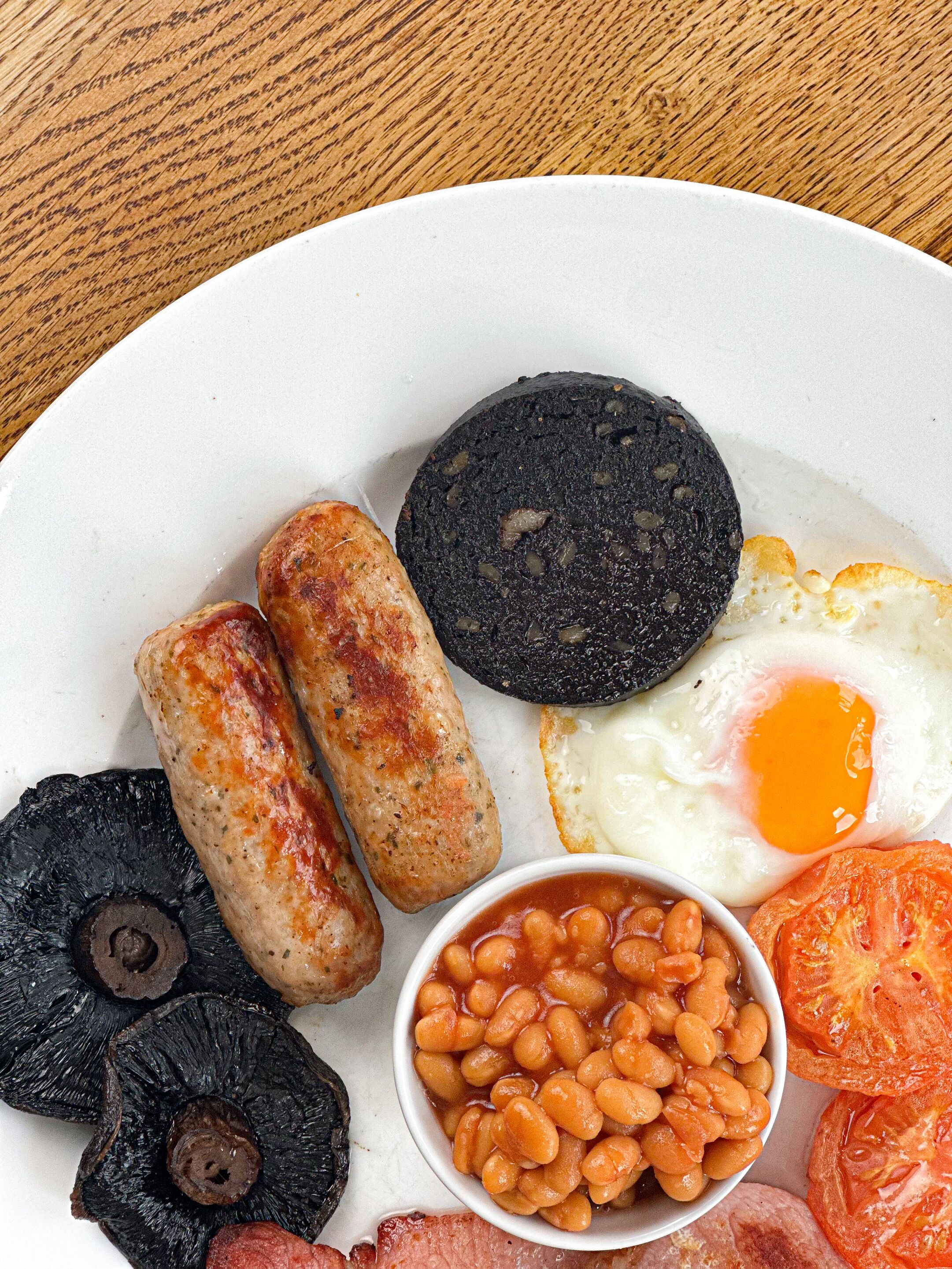 A plate of breakfast food including sausages, black pudding, fried egg, baked beans, and tomatoes.