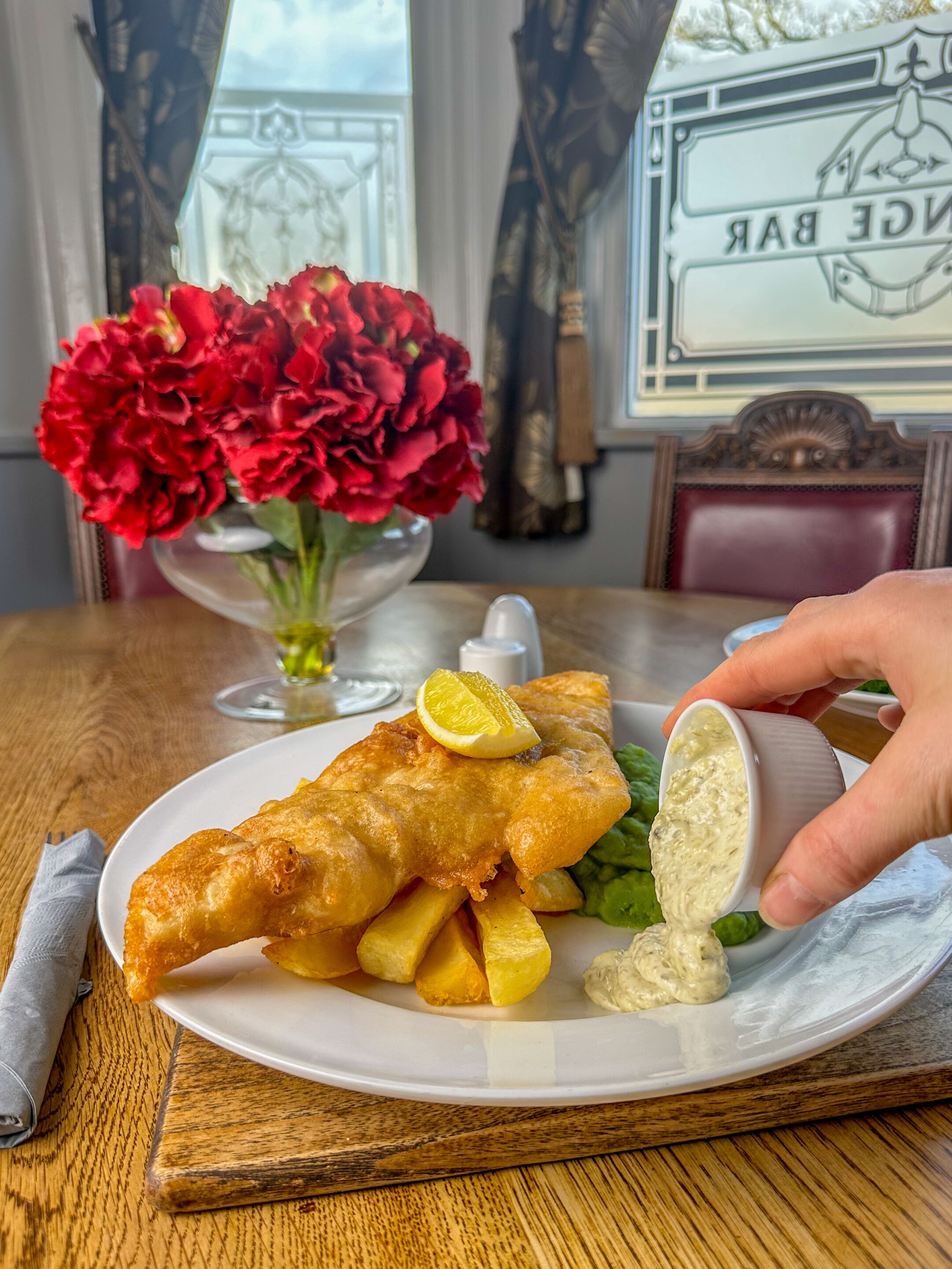 A plate of fish and chips with lemon wedges and sauce, a vase of red flowers, and a framed picture on the table.