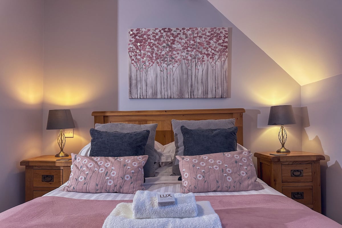 Ein ordentlich gemachtes Bett mit einer rosa Decke, zwei Kissen und einem gefalteten Handtuch mit einem Markenetikett. Es gibt zwei Nachttische mit Lampen, ein Holz-Kopfteil und ein Gemälde an der Wand.
