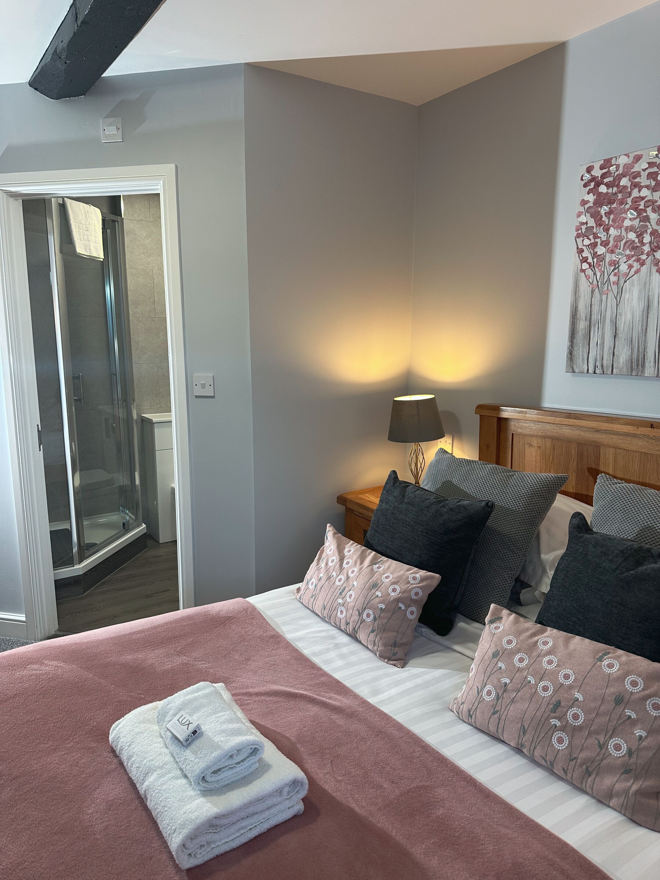 A hotel room with a bed, a nightstand, a lamp, a painting, and a sliding glass door leading to a shower area.