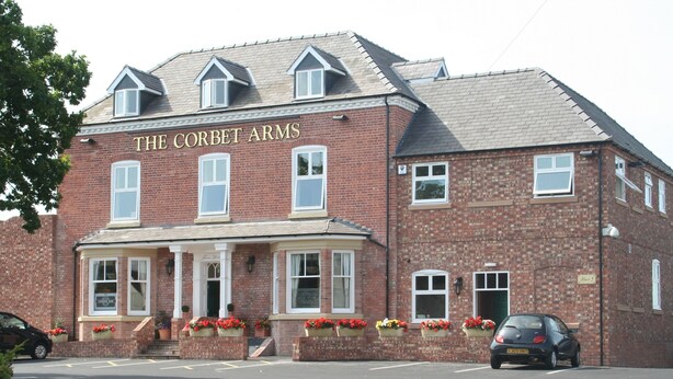 Ein zweistöckiges Backsteingebäude mit dem Schild "The Corbet Arms" über dem Eingang. Blumenkörbe stehen auf den Stufen und ein Auto ist vor der Fassade geparkt.