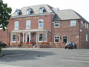 A two-story brick building with the sign "The Corbet Arms" above the entrance. There are flower baskets on the steps and a car parked in the front.