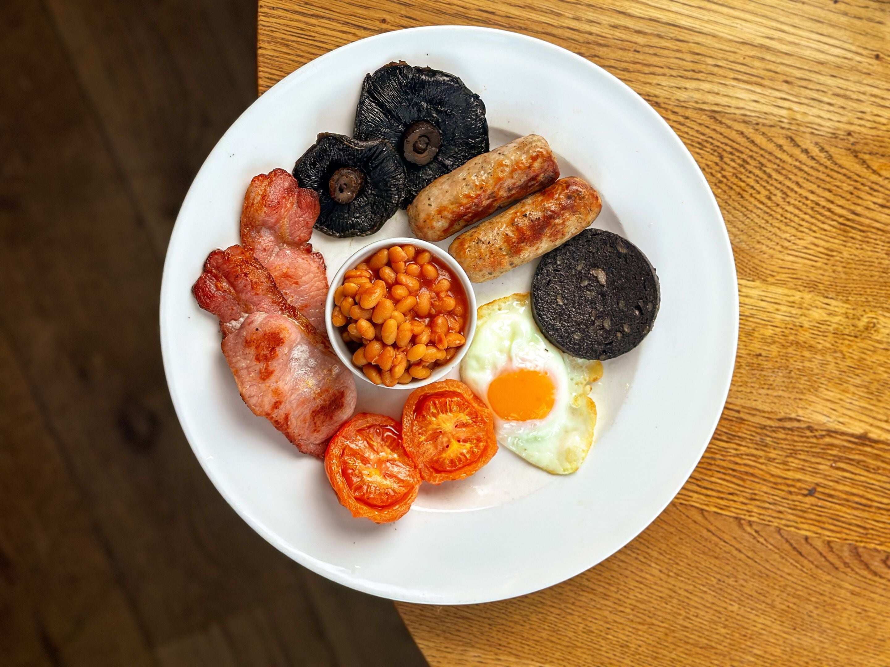 A plate of breakfast food including bacon, sausages, eggs, tomatoes, baked beans, and black pudding.