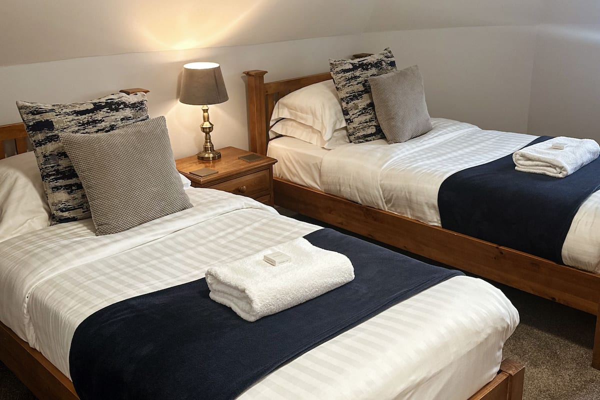 Ein Zimmer mit zwei hölzernen Betten, jeweils mit weißer, gestreifter Bettdecke, einer dunkelblauen Decke und einem weißen Handtuch. Ein Nachttisch mit Lampe und ein kleiner Tisch zwischen den Betten.
