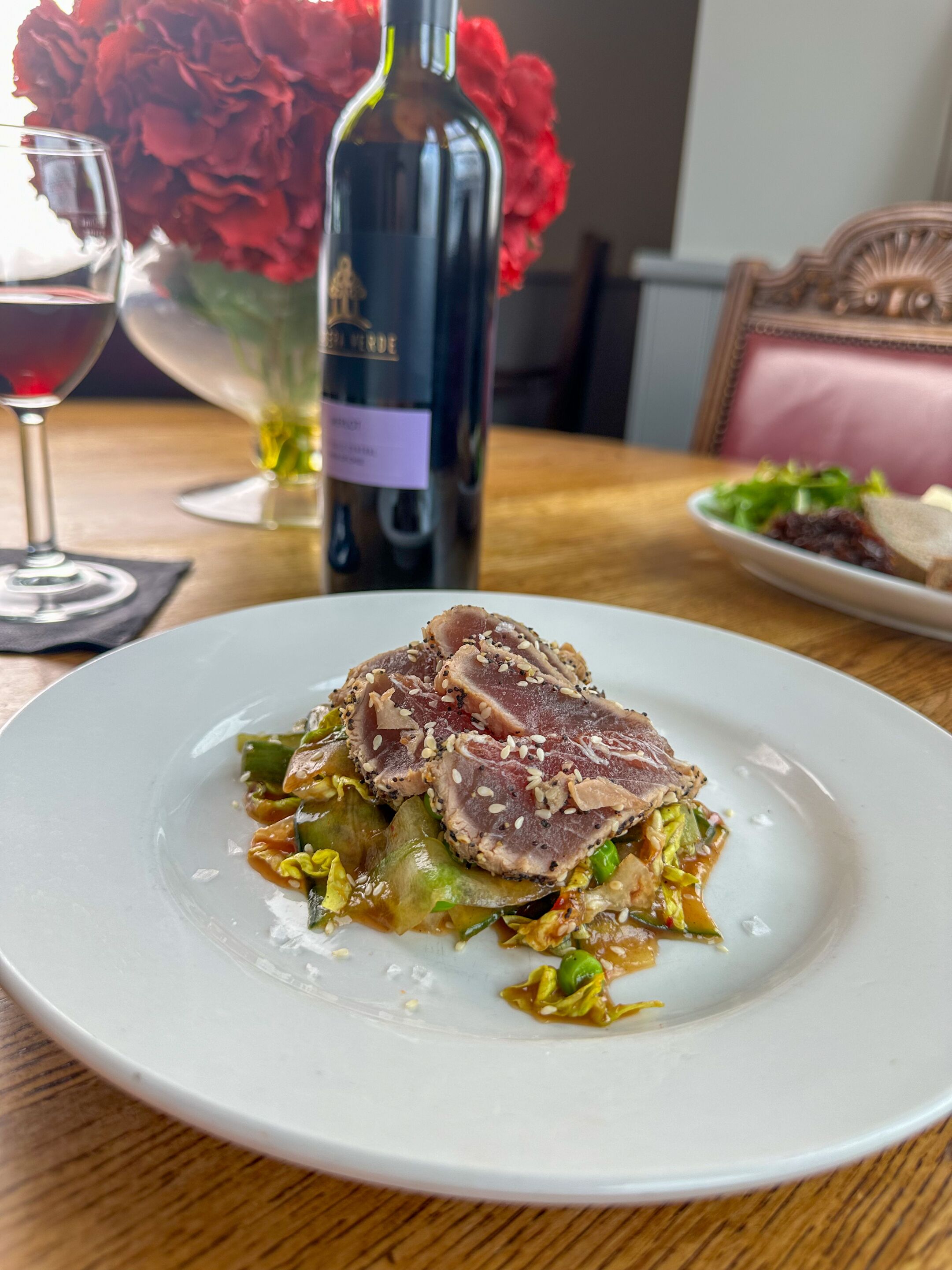 Un plato de comida con una botella de vino y flores al fondo.