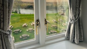 Ein Fenster mit zwei zurückgebundenen Vorhängen, das einen Blick auf eine Wiese mit einem Gewässer und Bäumen in der Ferne freigibt.