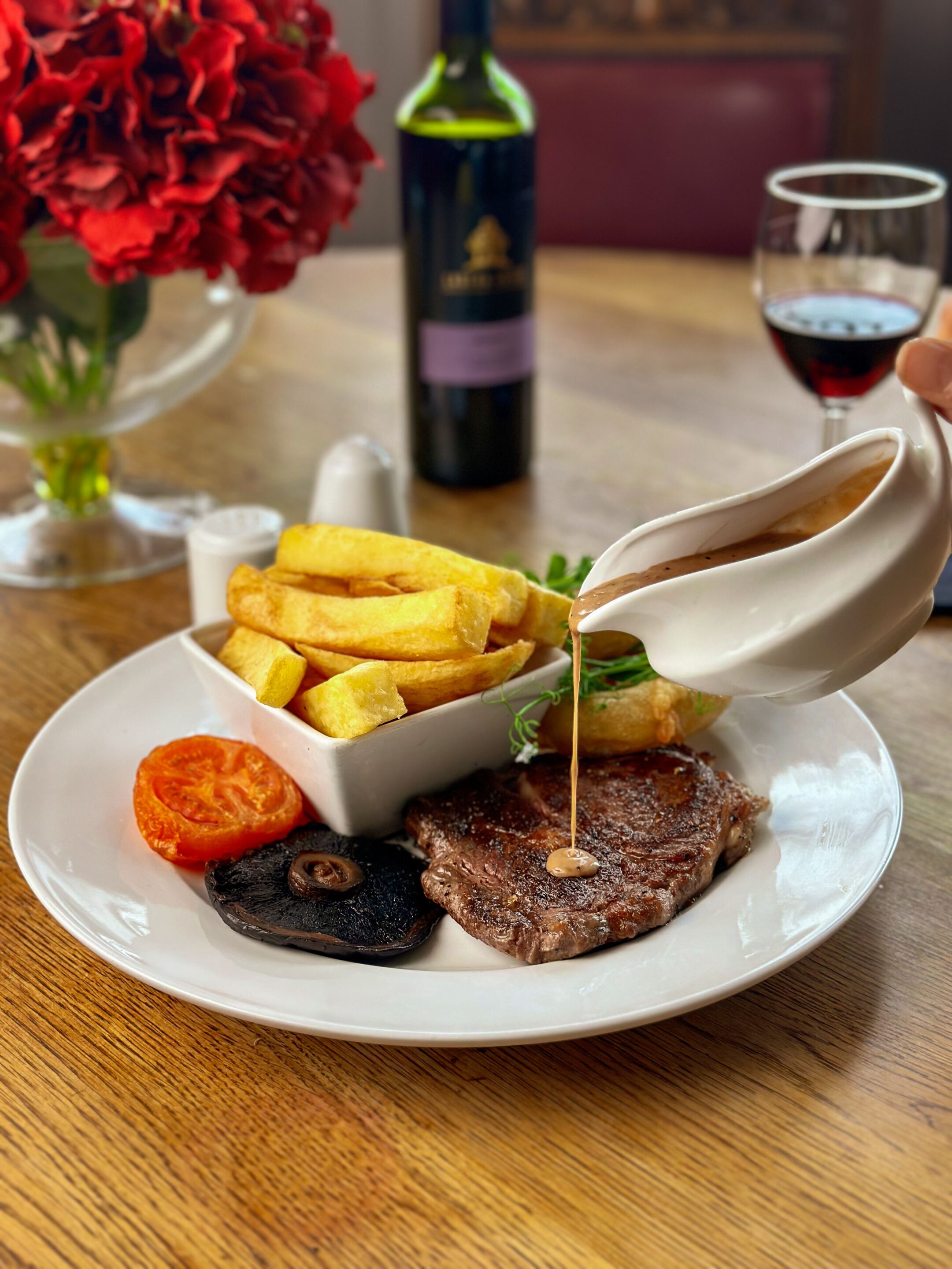 Un plato con bistec, papas fritas, una rodaja de tomate y champiñones, con salsa estando vertida sobre el bistec.