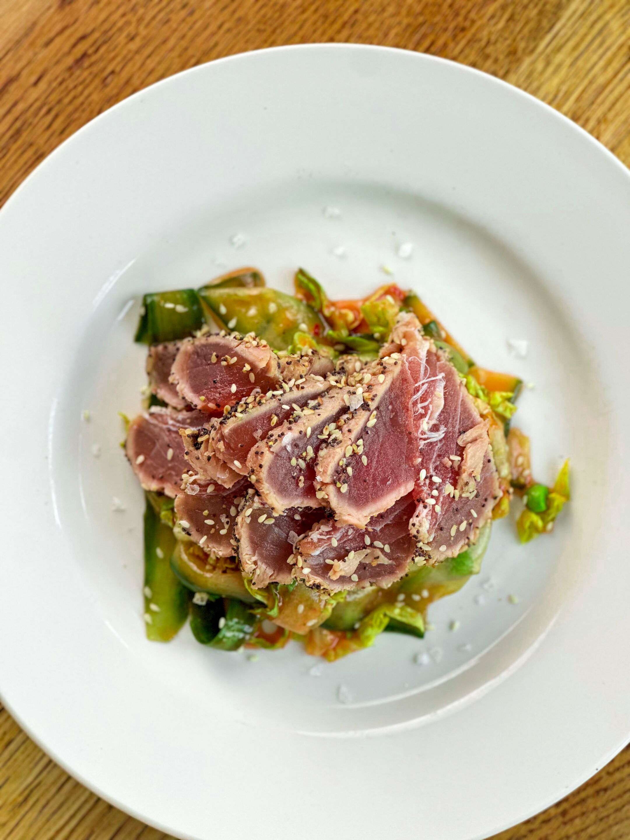 A white plate with a dish of sliced meat topped with sesame seeds, accompanied by a green vegetable salad.