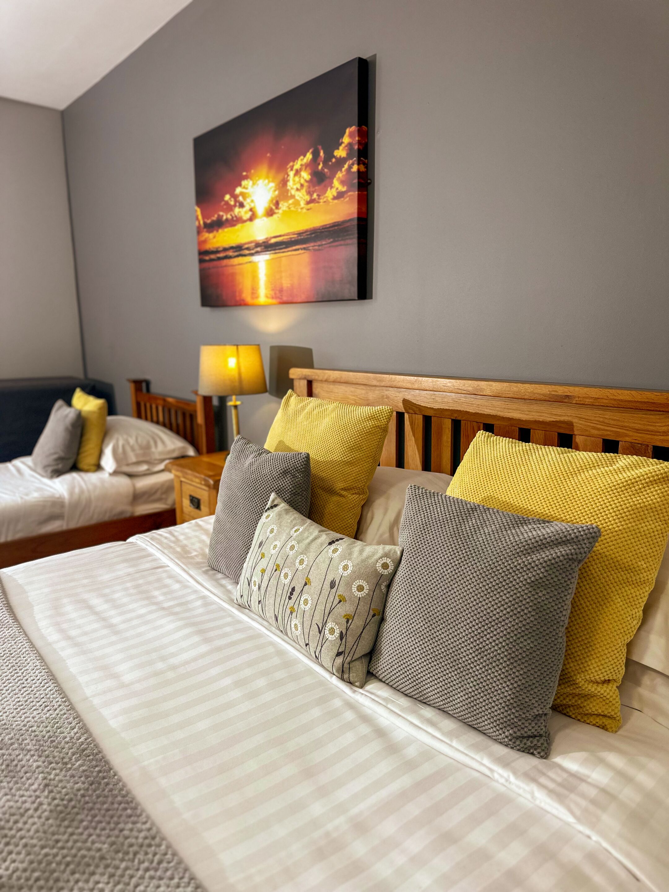 A neatly made bed with striped bedding and multiple pillows, a wooden headboard, and a wall-mounted painting of a sunset.