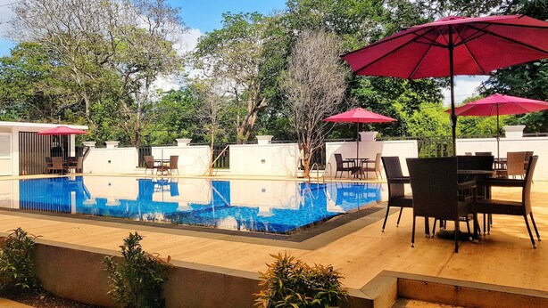 Una piscina con sillones de descanso y sombrillas, rodeada de árboles y un edificio blanco.