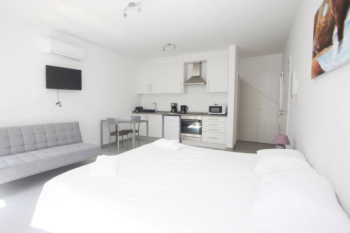 A modern, compact hotel room with a kitchenette, a sofa, a bed, and a TV mounted on the wall.