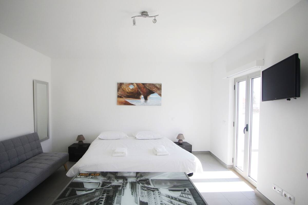 A modern hotel room with a bed, a gray sofa, a table with a cityscape print, a painting of a coastal scene, a ceiling fan, and a TV on the wall.