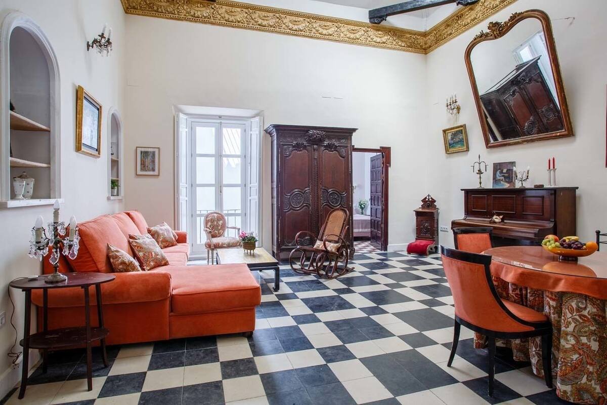 A room with a checkered floor, a sofa, a rocking chair, a dining table with a fruit bowl, and a large mirror.