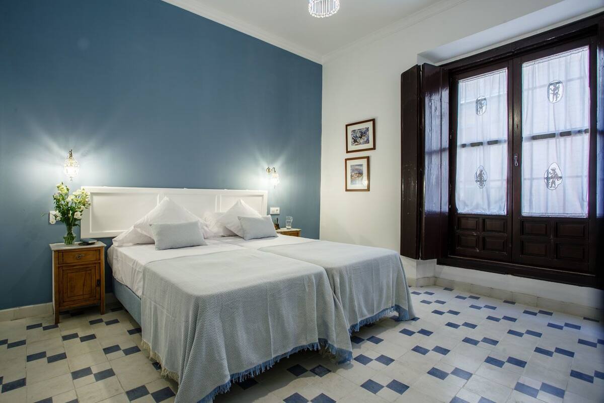 A bedroom with two beds, a nightstand with a vase of flowers, and a window with curtains.