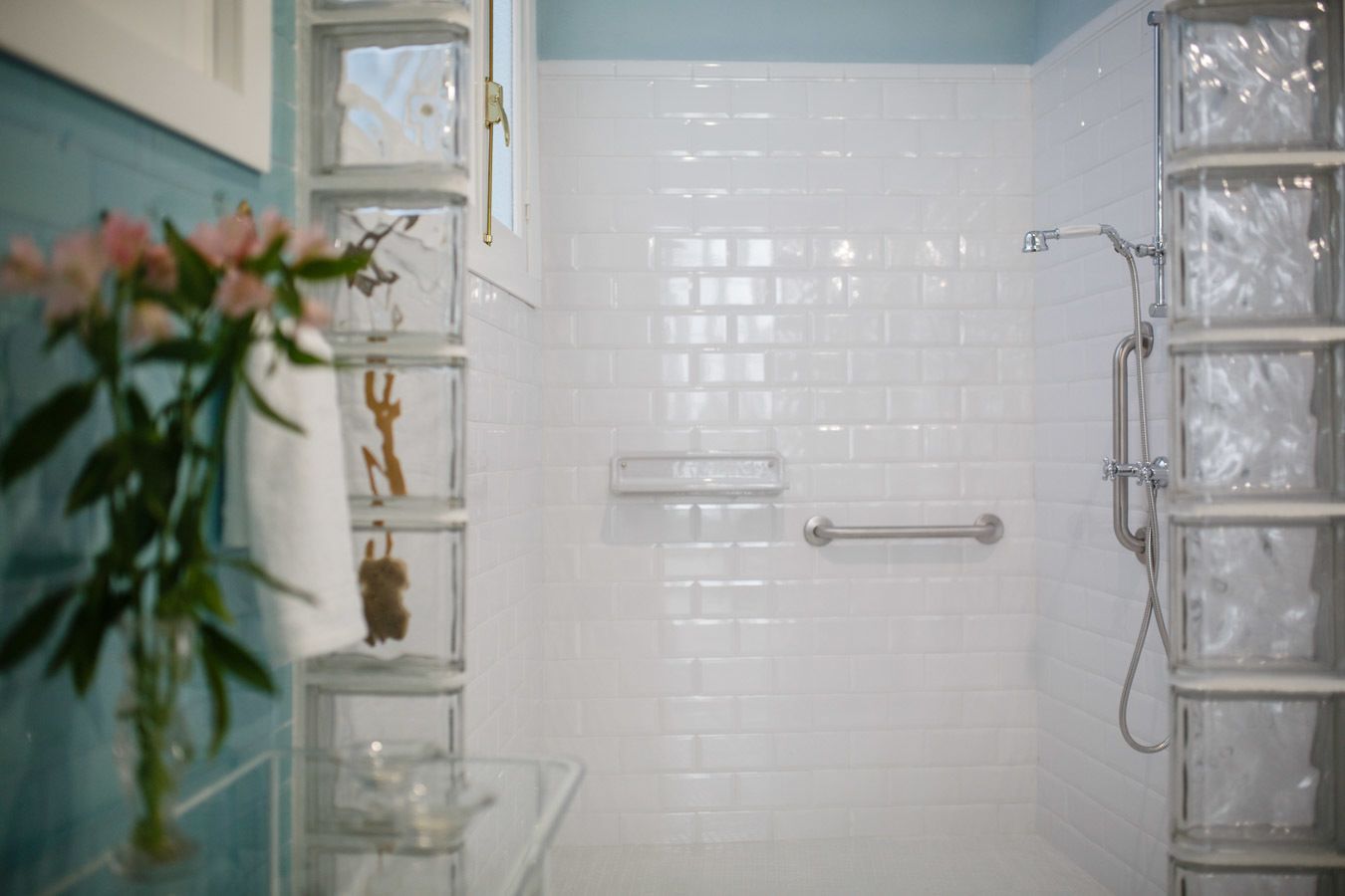 Une salle de bain avec des murs recouverts de carreaux blancs, une douche en verre et une étagère en verre transparent agrémentée d’objets décoratifs.