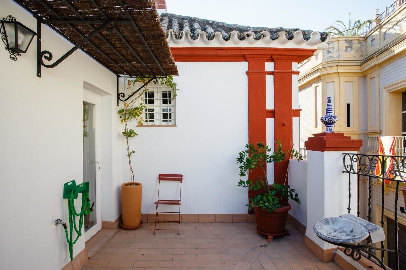 Une cour avec un toit en tuiles, une colonne rouge, des plantes en pot, une petite table et une chaise verte.