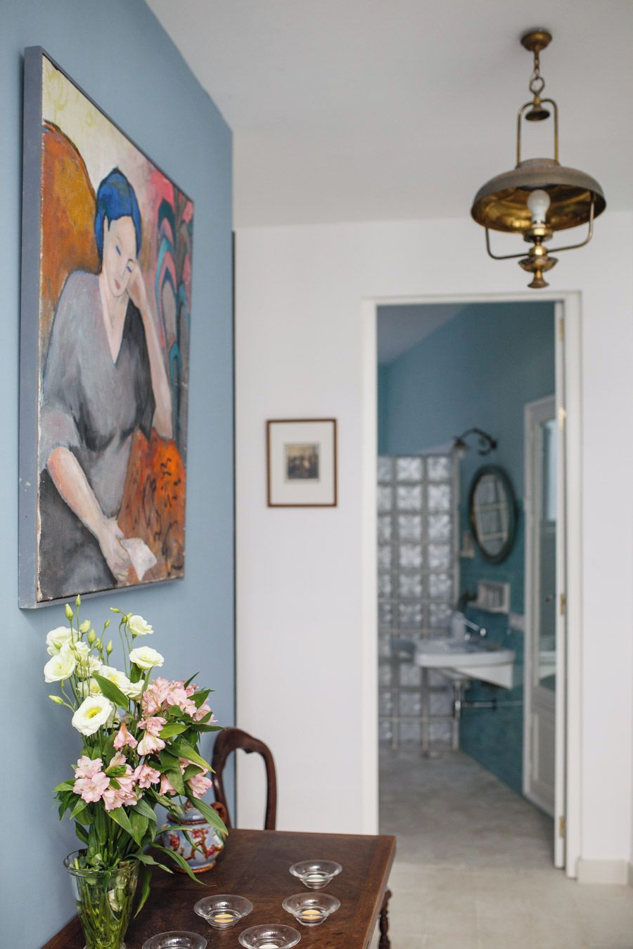 A dining area with a table, chairs, a vase with flowers, a painting on the wall, and a hallway leading to a bathroom.