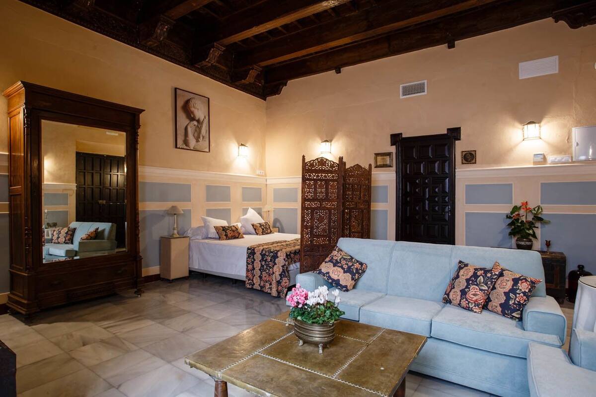 A room with a bed, a sofa, a coffee table with flowers, a mirror, and a wooden cabinet.