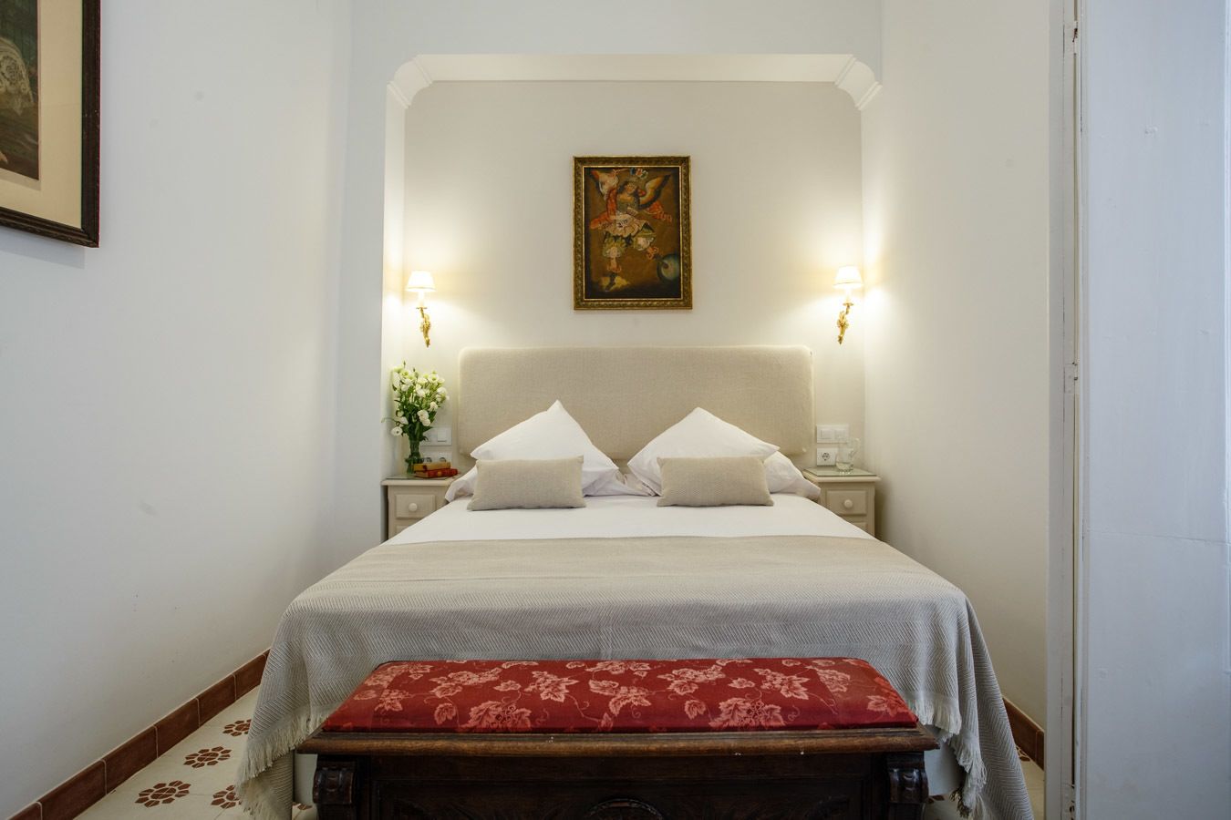 A hotel room with a bed, two bedside lamps, a painting on the wall, and a bench with a red cushion.