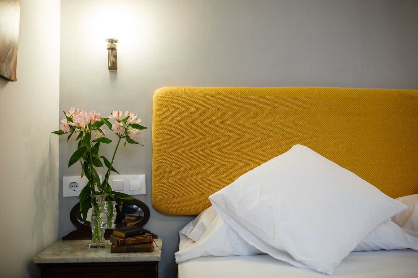 Un lit bien fait, avec des oreillers blancs et une tête de lit jaune, une table de chevet ornée d’un vase de fleurs et une lampe fixée au mur.