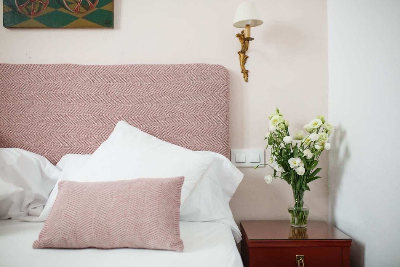 A neatly made bed with white and pink pillows, a wall-mounted lamp, and a vase with white flowers on a bedside table.