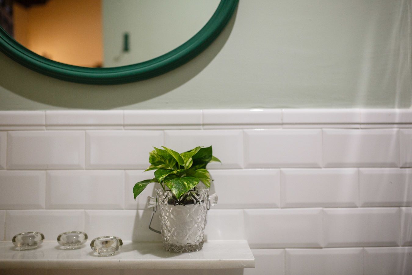 A bathroom with a white tiled wall, a green-framed mirror, and a small potted plant on a white shelf.
