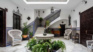 En rummelig gårdsplads med en central fontæne, omgivet af hvide fletstole og krukker med planter. En trappe med blå og hvide mønstrede gelændere fører til en etage over.