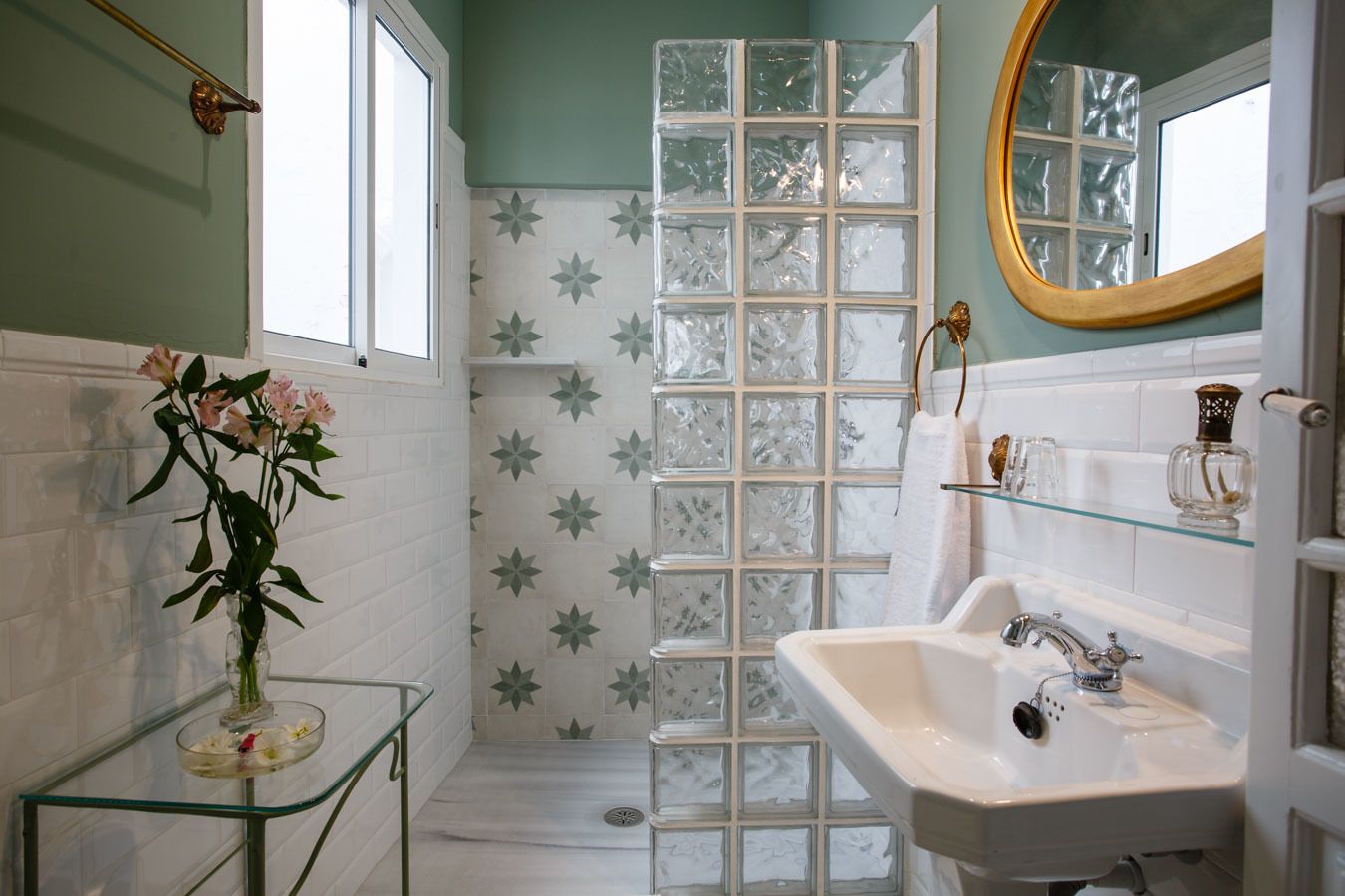 A bathroom with a glass shower enclosure, a white sink, and a vase with flowers on a glass table.