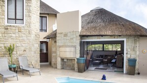 Uma área de piscina com espreguiçadeiras, uma construção com telhado de palha e um edifício de pedra.