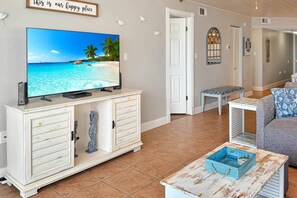 Un soggiorno con una televisione che mostra una spiaggia, un mobiletto bianco con cassetti, una panca e un divano a fantasia.