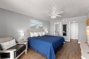 A bedroom with a large bed, a nightstand, a chair, a ceiling fan, and a painting on the wall.