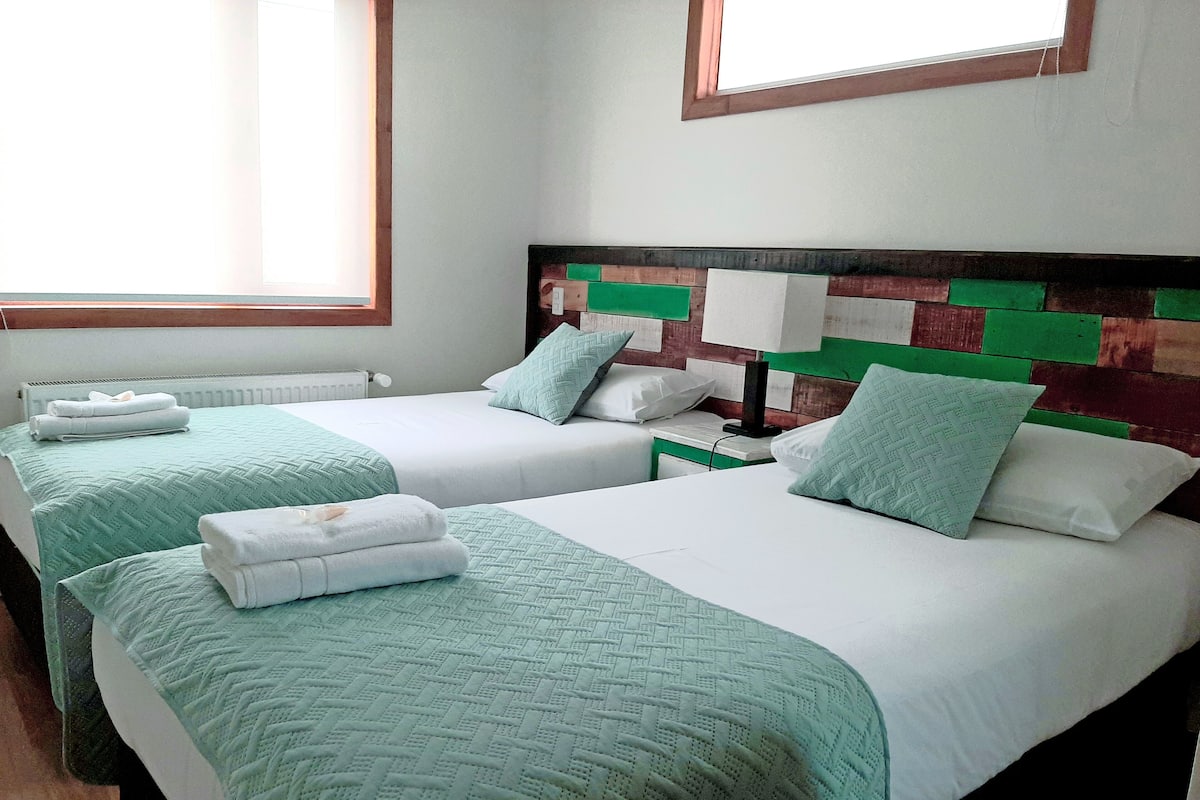 Ein Hotelzimmer mit zwei Betten, jedes mit einer grünen Steudecke und weißen Bettwäsche. Zwischen den Betten steht ein Nachttisch mit einer Lampe.